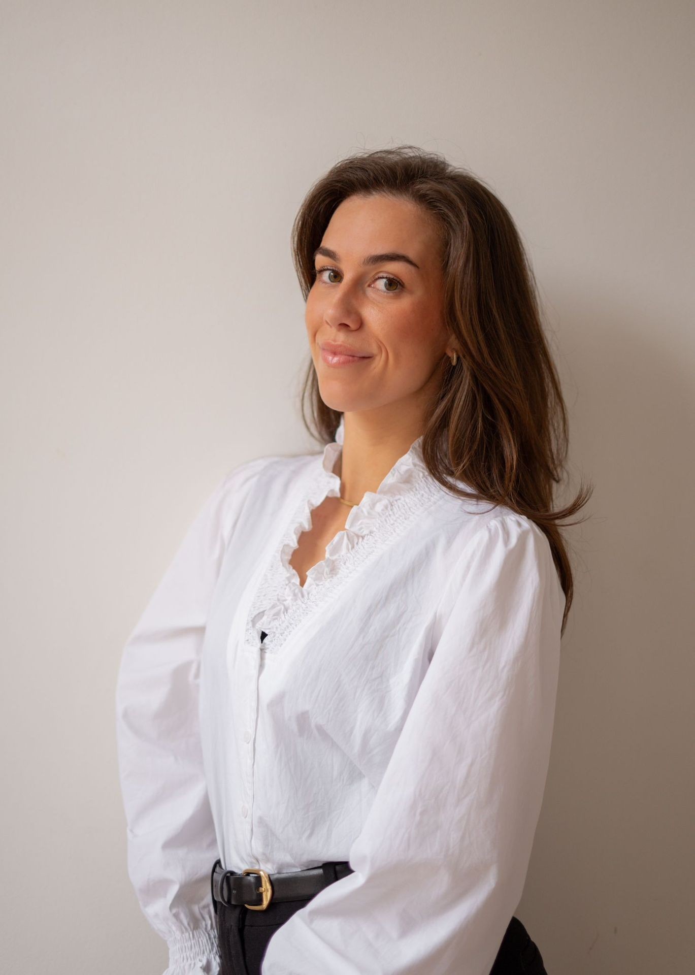 Person wearing a white blouse with ruffled collar and black belt, standing against a neutral background.